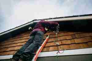 A person in a red flannel shirt standing on a ladder while carefully installing multicolored holiday light along the edge of a home's roof, showcasing the effort and precision needed for holiday lighting installation.